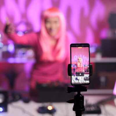 Woman with pink hair playing techno song at professional turntables while filming music process with phone camera, having fun in studio. Dj artist doing performance at nightclub with audio equipment
